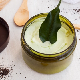 Słoik zielonego kremu z liściem na górze stoi na jasnej powierzchni, otoczony drewnianymi łyżkami, fusami z kawy, ręcznikiem i pokrojoną pomarańczą, tworząc naturalną, uspokajającą atmosferę spa.