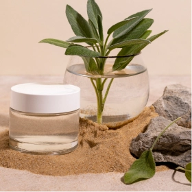 Szklany słoik z białą pokrywką stoi na piasku obok kamieni i wazonu z zielonymi liśćmi. Scena jest minimalistyczna i oddaje naturalny, spokojny nastrój. 