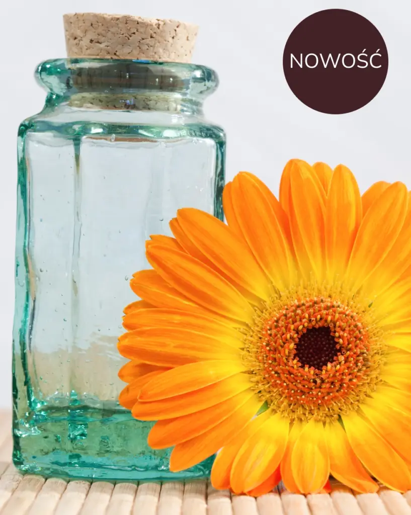 Przezroczysta szklana butelka z korkową pokrywką stoi obok intensywnie pomarańczowej gerbery na bambusowej macie. Zwinięte białe ręczniki sugerują atmosferę spa, przekazując spokój.