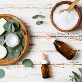 Drewniane miseczki z solą morską i produktami do pielęgnacji skóry spoczywają na liściach eukaliptusa. Dwie bursztynowe buteleczki z zakraplaczem dodają spa-podobnego charakteru białemu drewnu.